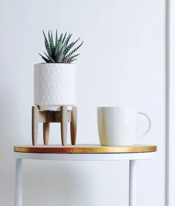 A white table with a gold top against a white wall. There is a potted plant and mug on top of the table.